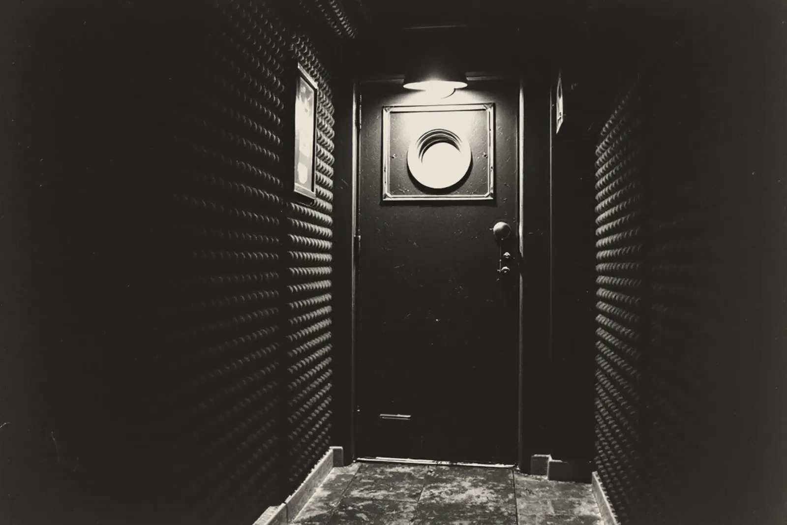A corridor lined with acoustic foam leading to a heavy studio door with a round porthole window, a single lamp over it.